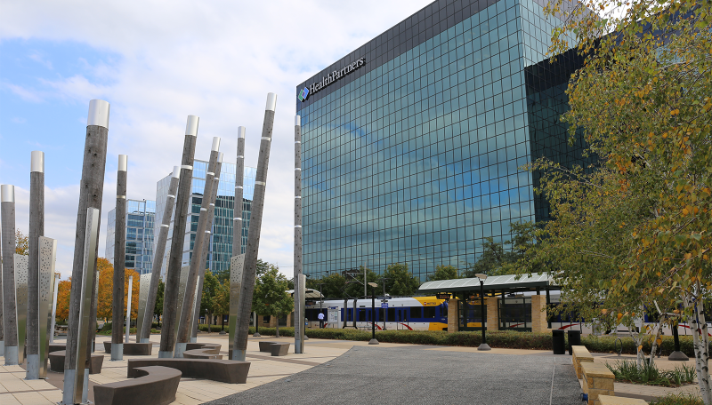 An urban park and light rail station in Bloomington, featuring an art installation in the foreground, office buildings in the background, and a light rail vehicle at the station.